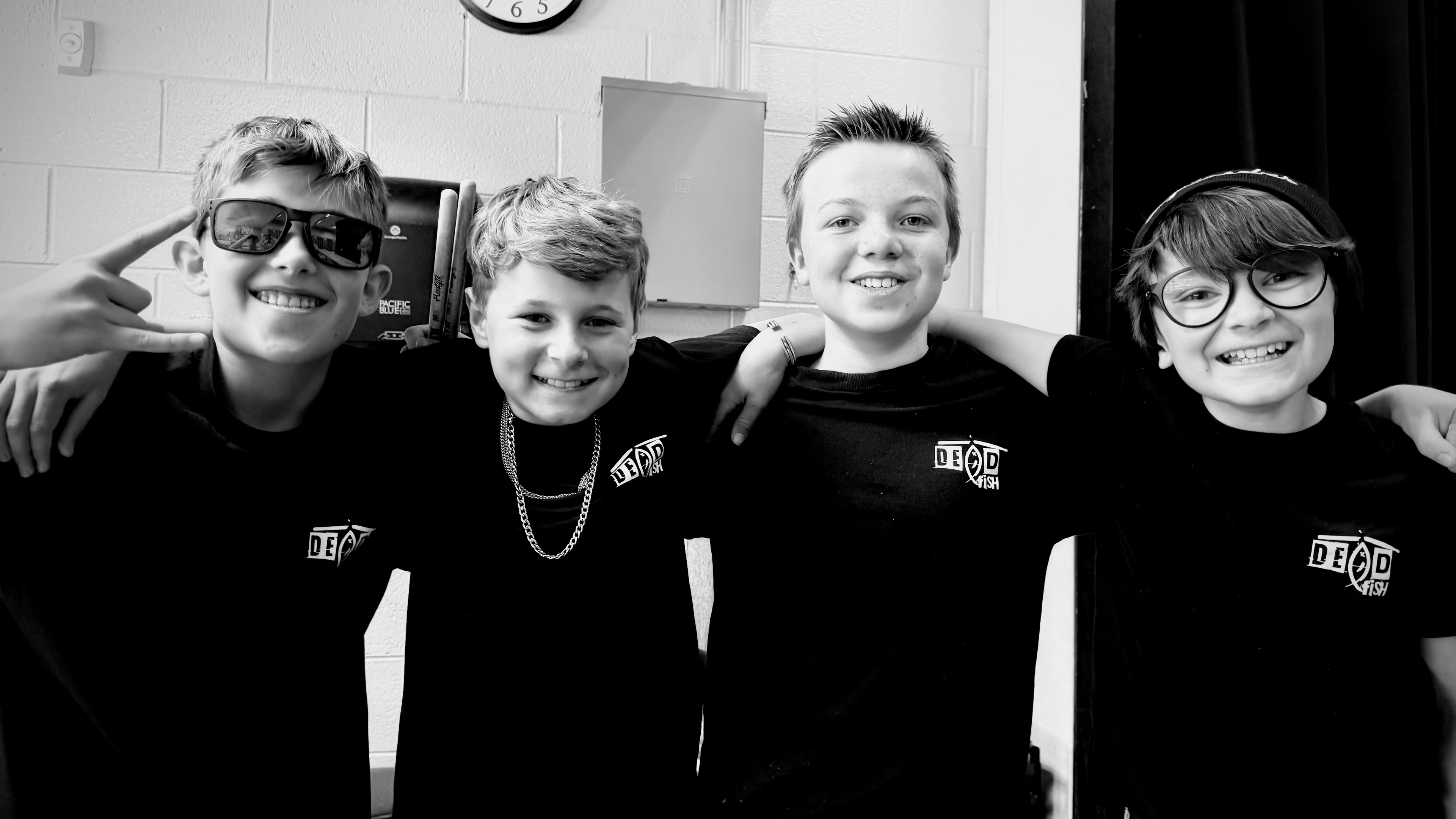 Four young people wearing matching black shirts with a logo, standing in a room with a clock on the wall.